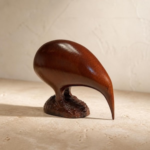 Peut inclure: Une sculpture en bois brun foncé représentant un kiwi. L'oiseau possède un corps arrondi et bulbeux ainsi qu'un long bec courbé vers le bas qui touche la base. La base est texturée et brun foncé, contrastant avec la finition polie du corps.