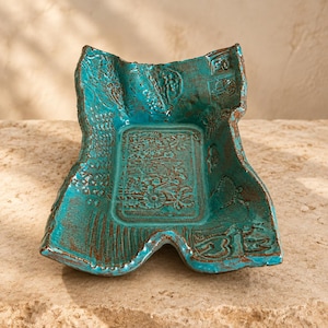May include: A rectangular ceramic tray with an irregular, wavy edge and a vibrant turquoise glaze. The surface features intricate, recessed floral and geometric patterns with dark brown accents highlighting the textured, stamped details.