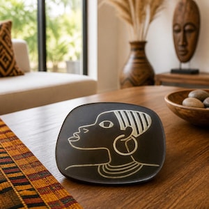 May include: A black decorative plate with a white etched profile of a person wearing a large hoop earring and multiple necklaces. The plate is on a wooden table with a colorful runner.