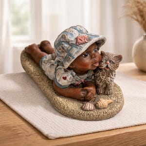 May include: A figurine of a child lying on a sandy surface, wearing a blue and white plaid hat and outfit, with a small kitten. The child has dark skin and is looking at the kitten. Seashells are on the sand.