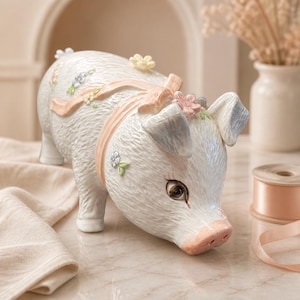 May include: A white ceramic piggy bank with a textured surface, featuring a soft peach-colored ribbon tied around its neck. The pig is decorated with small, delicate sculpted flowers in pastel shades of blue, yellow, and pink, and has a pink snout.