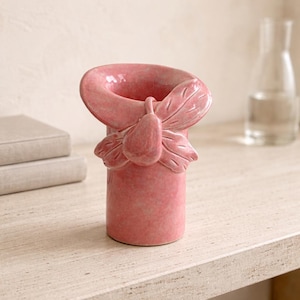 May include: A pink ceramic vase with a unique design. The vase features a cylindrical base and a flared top with a decorative leaf and pear-shaped accent. The vase is approximately 15 cm tall.