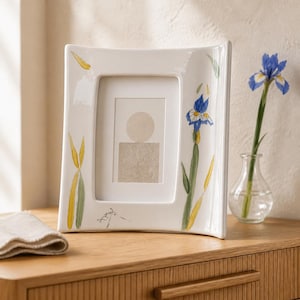 May include: A white ceramic picture frame with a curved, concave silhouette. The frame features hand-painted botanical illustrations of blue iris flowers and yellow leaves. A small black signature is visible on the lower left side of the frame.