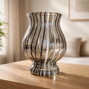 May include: A clear glass vase with a bulbous shape and a flared top. The vase is adorned with vertical stripes in black and white. The vase sits on a wooden surface, suggesting a decorative home accent.