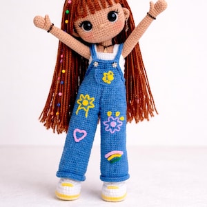 Peut inclure: Poupée au crochet faite à la main, avec de longs cheveux roux et une salopette en jean bleue décorée de fleurs brodées colorées, d'un cœur et d'un arc-en-ciel. La poupée porte des baskets blanches à semelles jaunes et lève les bras.