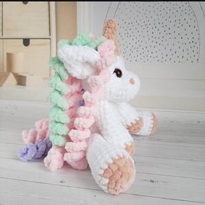 Peut inclure: Une licorne blanche faite à la main au crochet, avec une crinière et une queue aux couleurs pastel arc-en-ciel. La licorne a une corne et des sabots beiges, et des yeux bruns. Le jouet en peluche est posé sur une surface en bois clair.