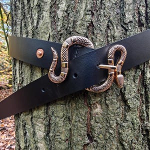 May include: Black leather belt featuring a bronze snake buckle. The belt is wrapped around a tree trunk, highlighting the intricate design of the snake clasp and the leather's texture. A copper rivet is also visible.
