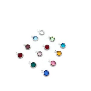 Pode incluir: Conjunto de 12 pequenos pingentes redondos com cristais de diferentes cores. Cada pingente tem um la&ccedil;o prateado para prender a um bracelete ou colar.