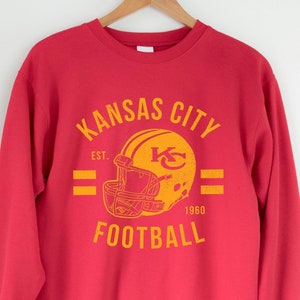 Pode incluir: Camisola vermelha com um gr&aacute;fico de estilo vintage de um capacete de futebol e o texto "Kansas City Football Est. 1960".