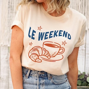 Może przedstawiać: Beżowa koszulka z niebieskim napisem "Le Weekend", wraz z brązową grafiką rogalika i filiżanki kawy. Osoba nosi niebieskie dżinsy.