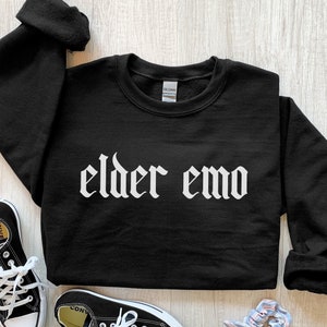 Puede incluir: Sudadera negra con el texto "elder emo" en fuente gótica blanca.