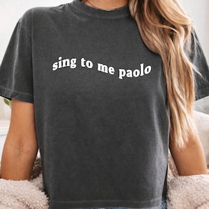 May include: A charcoal gray short-sleeve t-shirt with a crew neckline. The shirt features the white text "sing to me paolo" printed in a slightly curved, lowercase font across the chest. The fabric has a washed, vintage-style texture.
