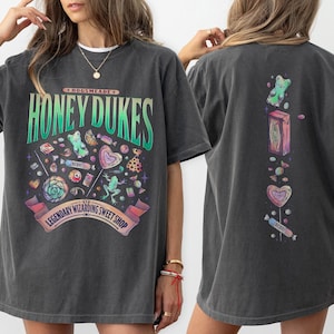 Può includere: Una maglietta grigia scura con una stampa grafica colorata di caramelle e il testo "Hogsmeade Honey Dukes Legendary Wizarding Sweet Shop".