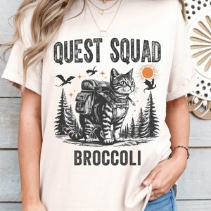 Könnte beinhalten: Ein beiges T-Shirt mit einer Schwarzweißillustration einer Katze, die einen Rucksack trägt und durch einen Wald geht. Der Text "QUEST SQUAD BROCCOLI" ist auf das T-Shirt gedruckt.