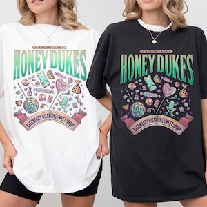 Może przedstawiać: Białe i czarne koszulki z logo Honeydukes, wzorem sklepu ze słodyczami z cukierkami, lizakami i żabą. Tekst brzmi "Hogsmeade Honeydukes Legendary Wizarding Sweet Shop".