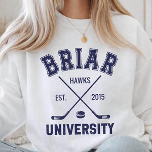 May include: White sweatshirt with a navy blue design featuring the word "BRIAR" above "HAWKS", crossed hockey sticks, a puck, and the text "EST. 2015" and "UNIVERSITY".