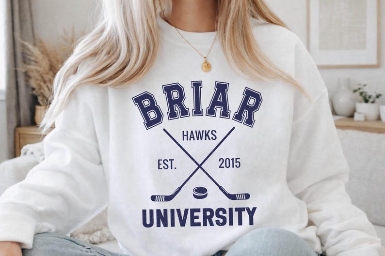 May include: White sweatshirt with a navy blue design featuring the word "BRIAR" above "HAWKS", crossed hockey sticks, a puck, and the text "EST. 2015" and "UNIVERSITY".