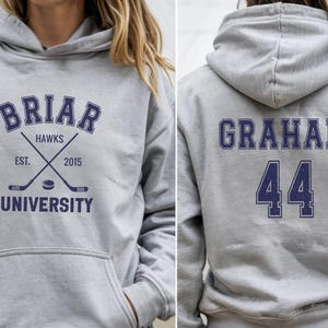 May include: A light gray hoodie with a front graphic that reads "Briar Hawks University" with crossed hockey sticks and a puck. The back of the hoodie displays the name "Graham" and the number "44" in navy blue.