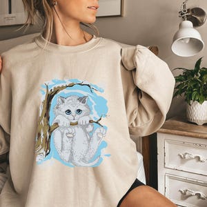 Pode incluir: Camisola bege com um gráfico de um gato branco com olhos azuis, empoleirado num galho sobre um fundo azul claro. O gato está segurando o galho. A camisola tem mangas compridas.