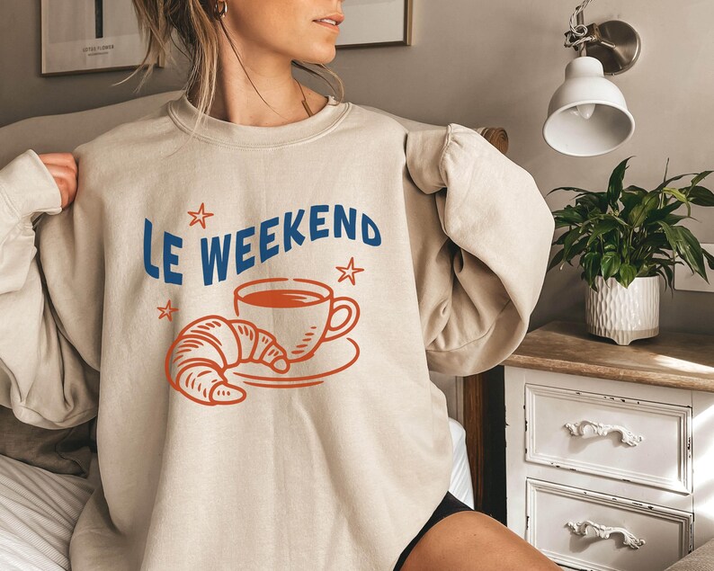 Może przedstawiać: Beżowa bluza z niebieskim napisem "Le Weekend", z czerwoną ilustracją filiżanki kawy, rogalika i gwiazdek. Bluza ma długie rękawy i okrągły dekolt.