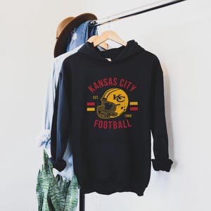 Pode incluir: Hoodie preto com um logotipo vintage do Kansas City Football com um capacete e o texto "Kansas City Football Est. 1960".