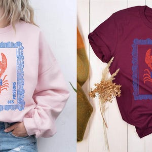 May include: Pink sweatshirt and burgundy t-shirt, both featuring a red lobster design within a blue frame. The text "LES POISSONS" is printed on the design. The sweatshirt is paired with blue jeans.
