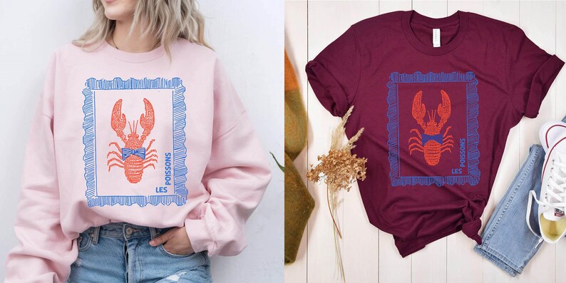 May include: Pink sweatshirt and burgundy t-shirt, both featuring a red lobster design within a blue frame. The text "LES POISSONS" is printed on the design. The sweatshirt is paired with blue jeans.