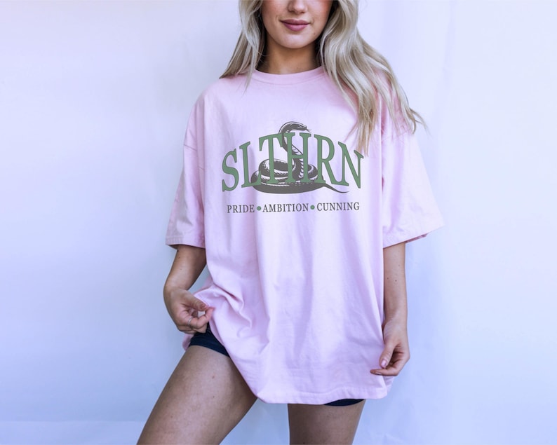 May include: A pink oversized t-shirt with a green snake graphic and the text "SLITHERN" with the words "PRIDE AMBITION CUNNING" underneath.