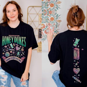 Może przedstawiać: Czarny t-shirt z napisem "Honeydukes" i grafiką cukierk&oacute;w. Prz&oacute;d przedstawia kolorowy wz&oacute;r r&oacute;żnych słodyczy, a tył ilustracje żelk&oacute;w i cukierk&oacute;w. Koszulka jest noszona przez osobę w niebieskich dżinsach.