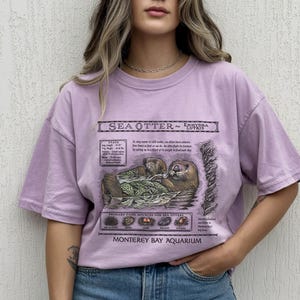 May include: A lilac-colored t-shirt featuring a detailed illustration of a sea otter, with scientific information and the text "Monterey Bay Aquarium." The graphic includes the sea otter's habitat and primary food sources.