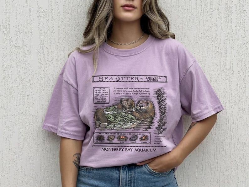 May include: A lilac-colored t-shirt featuring a detailed illustration of a sea otter, with scientific information and the text "Monterey Bay Aquarium." The graphic includes the sea otter's habitat and primary food sources.