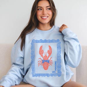 May include: Light blue sweatshirt featuring a red lobster illustration within a blue frame. The lobster has a blue bow tie. The words "LES POISSONS" are printed in blue on the right side of the frame. The sweatshirt is a casual style.