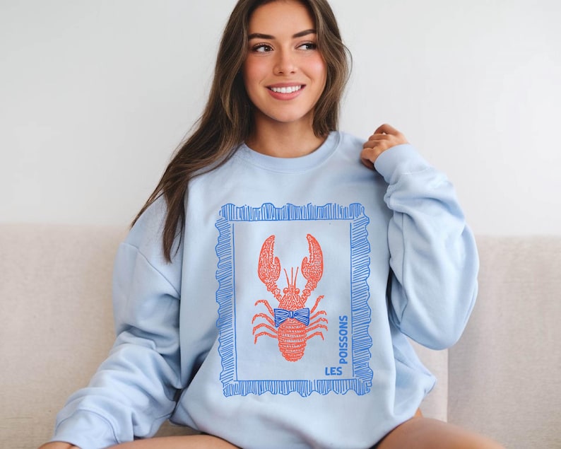 May include: Light blue sweatshirt featuring a red lobster illustration within a blue frame. The lobster has a blue bow tie. The words "LES POISSONS" are printed in blue on the right side of the frame. The sweatshirt is a casual style.