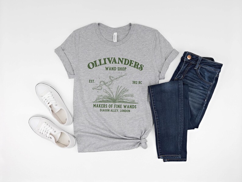 May include: A gray t-shirt with a green graphic of Ollivanders Wand Shop. The graphic includes the text "Ollivanders Wand Shop", "Est. 382 BC", and "Makers of Fine Wands Diagon Alley, London".