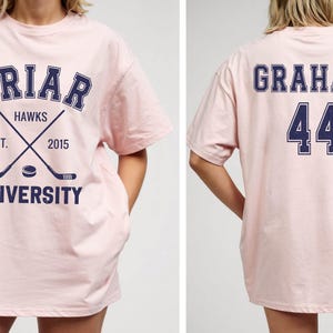 May include: A light pink t-shirt with the words "BRIAR UNIVERSITY HAWKS" in navy blue. The front of the shirt features crossed hockey sticks and a hockey puck. The back of the shirt displays the name "GRAHAM" and the number "44" in navy blue.