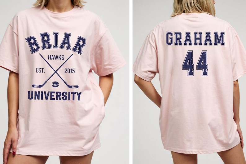 May include: A light pink t-shirt with the words "BRIAR UNIVERSITY HAWKS" in navy blue. The front of the shirt features crossed hockey sticks and a hockey puck. The back of the shirt displays the name "GRAHAM" and the number "44" in navy blue.