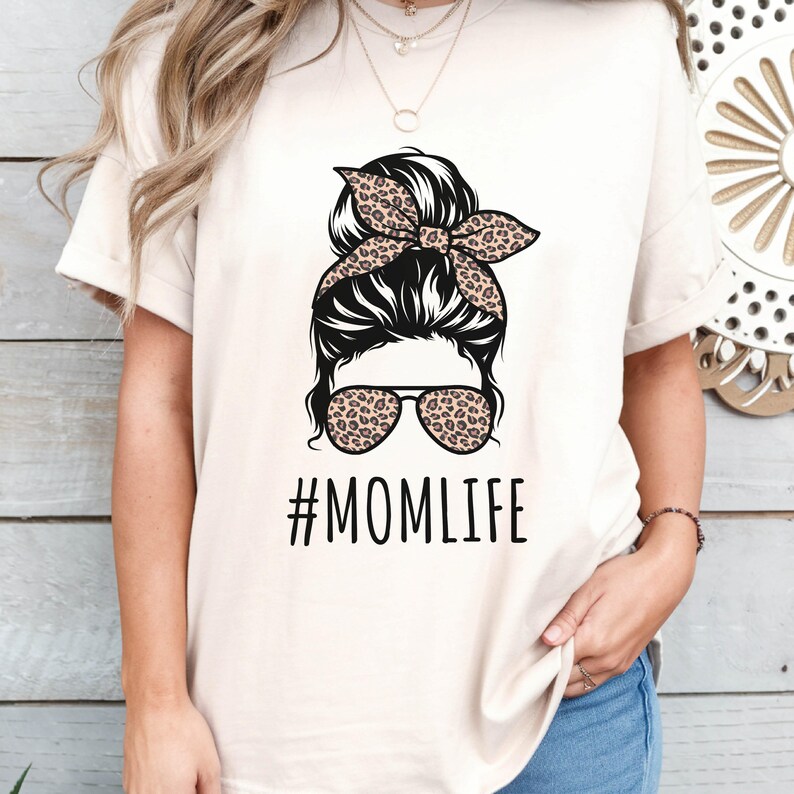 May include: A white t-shirt with a graphic of a woman with a messy bun, leopard print headband, and sunglasses. The text "#MOMLIFE" is printed below the graphic.