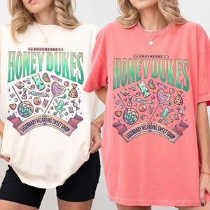 Może przedstawiać: Dwie koszulki, jedna w kolorze kości słoniowej, druga w kolorze arbuza, z logo Honeydukes. Projekt zawiera ilustracje cukierk&oacute;w i napis "Legendary Wizarding Sweet Shop". Koszulki mają kr&oacute;tkie rękawy i luźny kr&oacute;j.