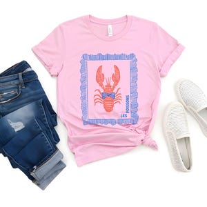 May include: Pink t-shirt with a red lobster graphic inside a blue frame. The shirt is paired with blue jeans and white slip-on shoes. The text "LES POISSONS" is printed on the shirt.