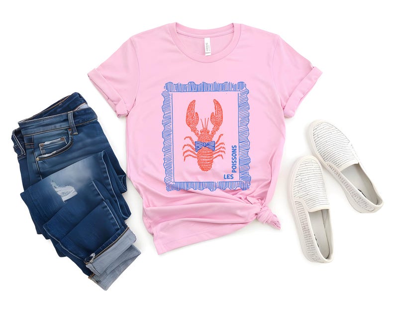May include: Pink t-shirt with a red lobster graphic inside a blue frame. The shirt is paired with blue jeans and white slip-on shoes. The text "LES POISSONS" is printed on the shirt.