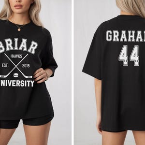 May include: Black t-shirt with white text and graphics. The front features "Briar Hawks University" with crossed hockey sticks and a puck. The back displays "Graham" and the number "44".