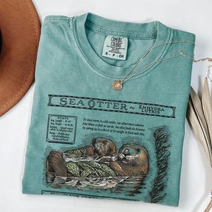May include: A teal Comfort Colors t-shirt featuring a detailed illustration of a sea otter with the text "SEA OTTER" and scientific information. A delicate silver necklace with a small pendant rests on the shirt.