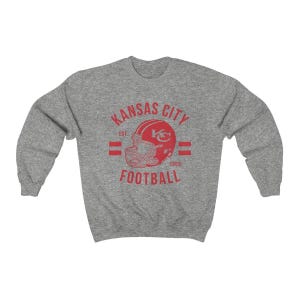 Pode incluir: Camisola cinzenta com um gr&aacute;fico vermelho de estilo vintage de um capacete de futebol com o texto "Kansas City Football Est 1960"