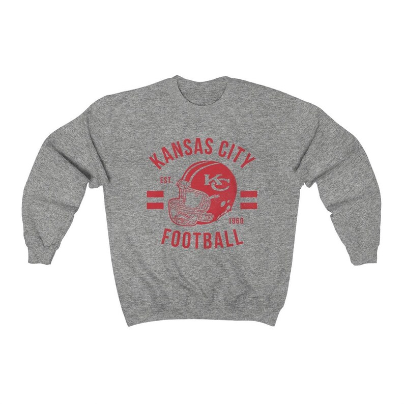 Pode incluir: Camisola cinzenta com um gr&aacute;fico vermelho de estilo vintage de um capacete de futebol com o texto "Kansas City Football Est 1960"