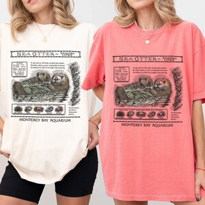 May include: Two short-sleeved t-shirts, one ivory and one watermelon, each featuring a detailed graphic of a sea otter with text that reads "Sea Otter - Enhydra Lutris" and "Monterey Bay Aquarium". The graphic includes illustrations of the otter and its food sources.