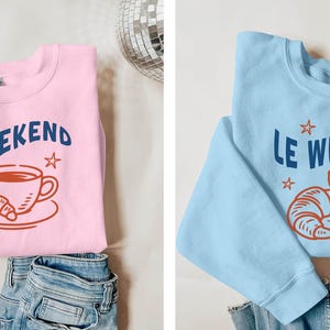 Może przedstawiać: R&oacute;żowa bluza z napisem "Le Weekend" w kolorze granatowym, wraz z grafiką filiżanki kawy i pomarańczowego rogalika. Bluza jest złożona na niebieskich dżinsach.