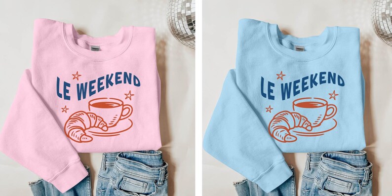 Może przedstawiać: R&oacute;żowa bluza z napisem "Le Weekend" w kolorze granatowym, wraz z grafiką filiżanki kawy i pomarańczowego rogalika. Bluza jest złożona na niebieskich dżinsach.
