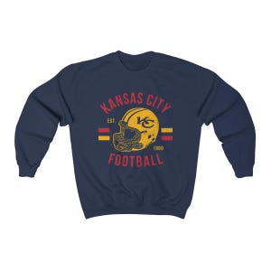 Pode incluir: Camisola azul-marinho com um logotipo vintage dos Kansas City Chiefs. O logotipo apresenta um capacete amarelo com um "KC" vermelho e o texto "Kansas City Football Est. 1960".