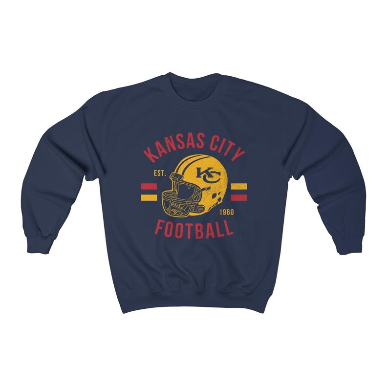 Pode incluir: Camisola azul-marinho com um logotipo vintage dos Kansas City Chiefs. O logotipo apresenta um capacete amarelo com um "KC" vermelho e o texto "Kansas City Football Est. 1960".