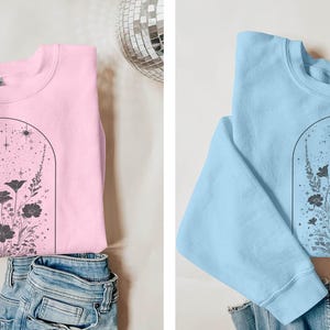 May include: Two sweatshirts with a floral design. One is pink and the other is light blue. Both have a black outline of a flower arrangement with a starry sky inside an arch.